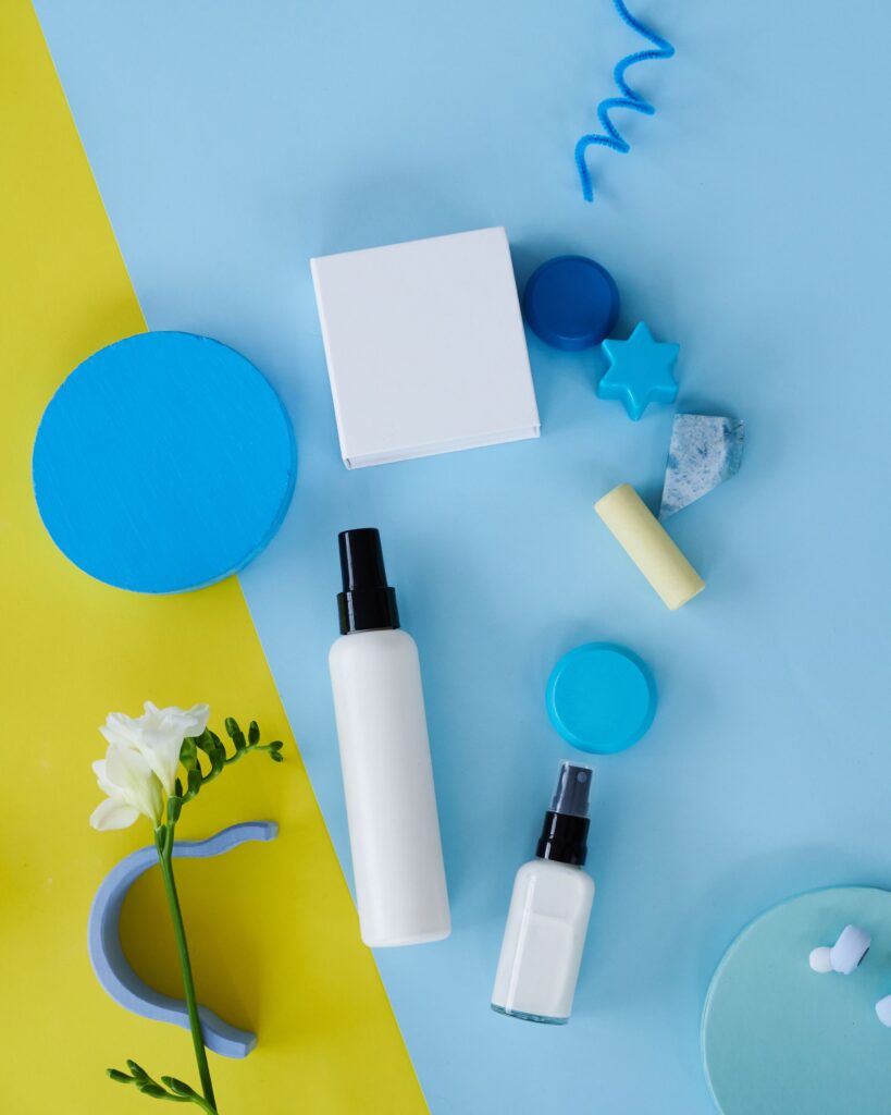 Flat lay view of skincare and cosmetic products, including white spray bottles, cream jars, and blue geometric display props arranged on a split yellow and light blue background, showcasing top beauty offers on sinibuy.com.